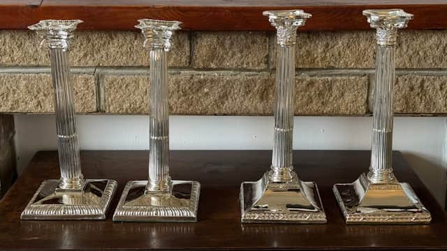 Four ornate silver candlesticks with fluted columns and square bases, displayed on a wooden surface against a stone wall.