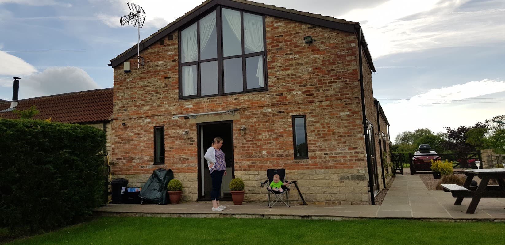 Rustic brick barn with large windows, a woman standing nearby, and a child sitting in a chair on a patio.