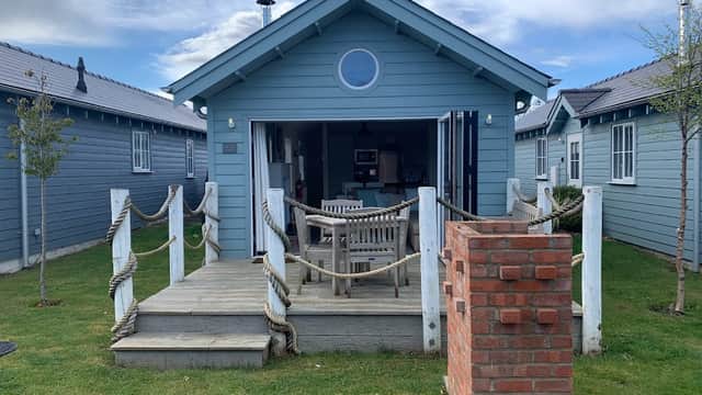 Blue beach house with a circular window, wooden deck, and brick barbecue, surrounded by green grass and other cabins.