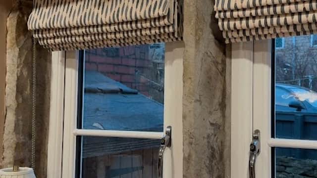 Wooden beams frame two windows adorned with patterned blinds, surrounded by decorative cards and a glass vase.