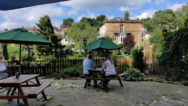 Two people sit at a picnic table under green umbrellas, surrounded by a garden and historic buildings in Plompton.