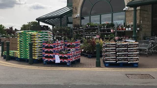 Morrisons store entrance with stacked pallets of plants, flags, and shopping carts in front under a cloudy sky.