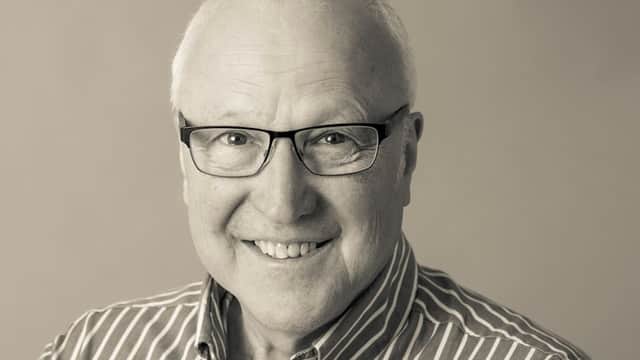 Smiling older man with glasses, wearing a striped shirt, against a neutral background.