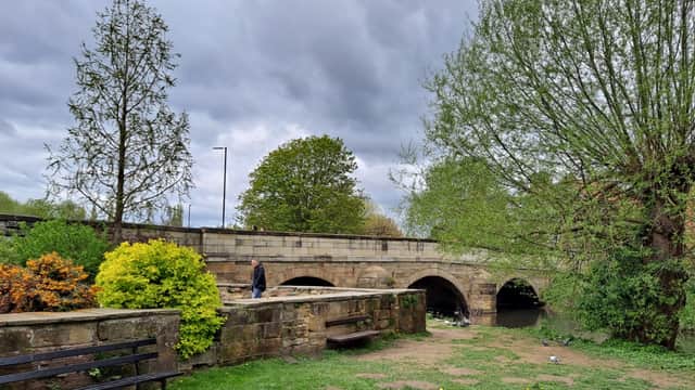 A stone bridge arches over a river, surrounded by lush greenery and colorful shrubs under a cloudy sky.