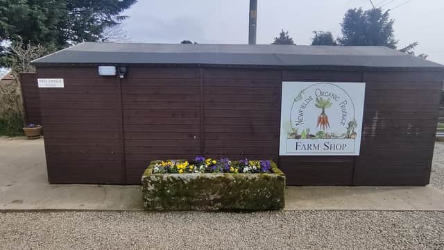 Wooden farm shop building with a sign reading "Newfields Organics" and a planter with colorful flowers at the entrance.