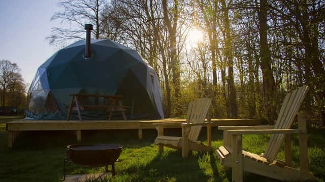 A geodesic dome structure with a chimney, surrounded by trees, wooden chairs, and a fire pit in a grassy area.