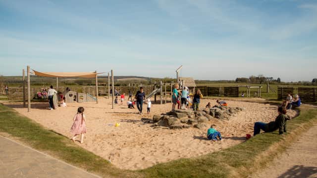 Children play on swings and a wooden structure, with sand below, in a rustic outdoor area at William's Den.