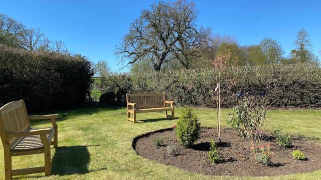A sunny community garden with wooden benches, a circular flower bed, and lush greenery in Buttercrambe, Yorkshire.