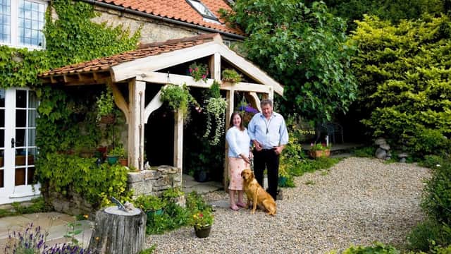 Charming bed and breakfast with a wooden entrance, lush greenery, a couple, and a dog in a gravel courtyard.
