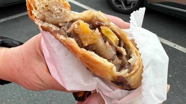 A hand holding a flaky pastry filled with meat and vegetables, with parked cars in the background.