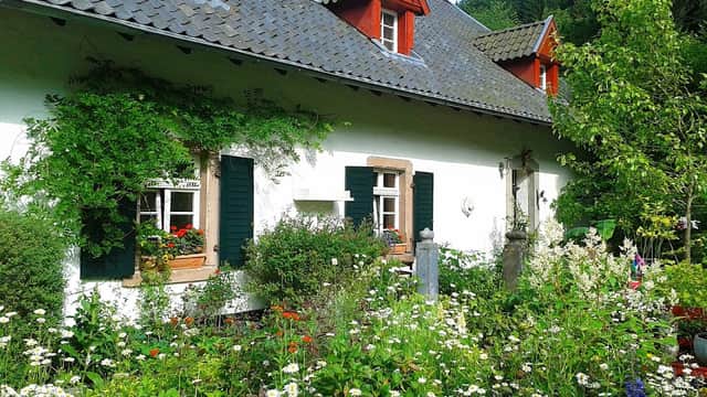 Charming farmhouse with red accents, surrounded by a lush garden filled with flowers and greenery.