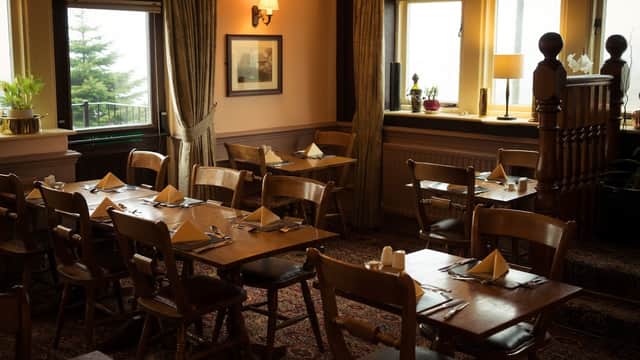Cozy dining area with wooden tables, chairs, and neatly set napkins, surrounded by warm lighting and large windows.