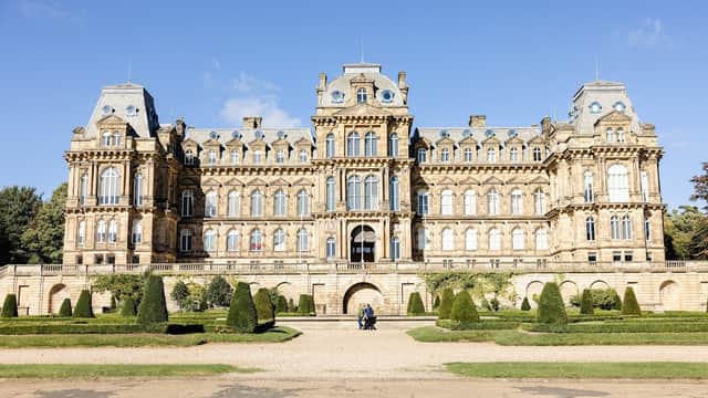 Victorian mansion with ornate architecture, surrounded by manicured gardens and trees under a clear blue sky.