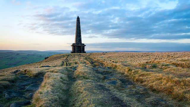 Tall stone monument atop grassy hill, surrounded by rolling hills and a cloudy sky at sunset.