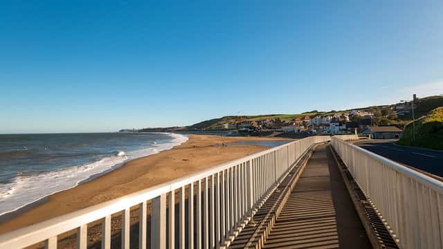 Coastal view from a walkway, with sandy beach, waves, and a quaint village in the background under a clear blue sky.
