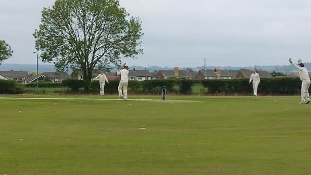 Cricket players in white uniforms on a grassy field, with trees and houses in the background under a cloudy sky.