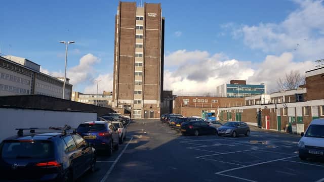 Rooftop car park with parked cars, a tall building in the background, and a clear blue sky with scattered clouds.