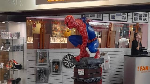 A colorful Spider-Man statue crouches on a brick pedestal in front of "The Fan Cave" shop, featuring memorabilia displays.