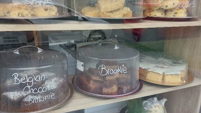 Display of assorted baked goods under glass domes, including scones, flapjack, brownies, and a teapot in the window.