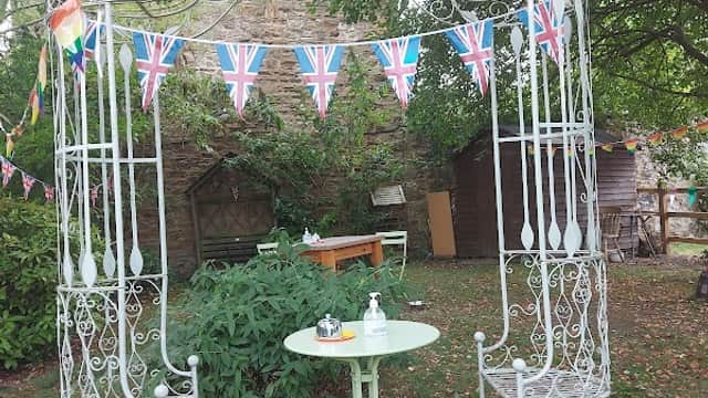 A quaint garden setting with a decorative gazebo, Union Jack bunting, and a small green table with chairs.