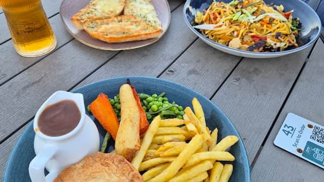 A table set with a pie, fries, peas, carrots, and salad, alongside drinks and garlic bread in a casual cafe setting.