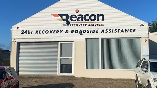 White building with "Beacon Recovery Services" sign, large windows, and gravel parking area. Clear blue sky above.
