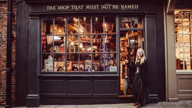 A woman in a long black dress stands outside a whimsical hobby shop with a large window displaying various colorful items.