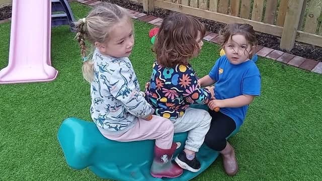 Three children sit on a green rocking toy in a playground, with a pink slide and wooden fence in the background.