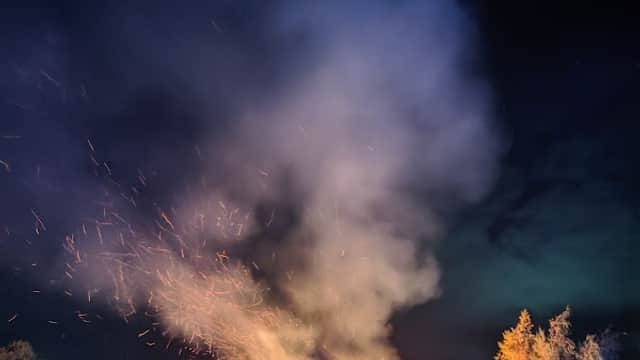 Crowd gathered around a large bonfire with smoke and sparks rising into the night sky, surrounded by trees.