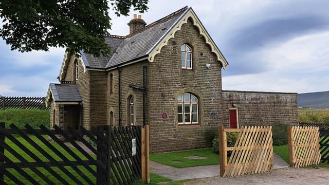 Historic stone building with a gabled roof, surrounded by a wooden fence and green grass, set against a cloudy sky.