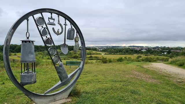 Metal sculpture featuring various tools, with a grassy landscape and cloudy sky in the background.