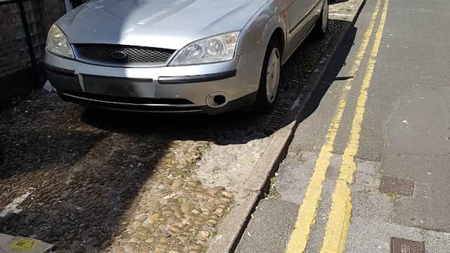 Silver car parked on a narrow street with yellow lines, surrounded by buildings and a clear blue sky.
