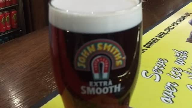 Pint of John Smith's Extra Smooth on a bar counter with drinks and promotional signage in the background.