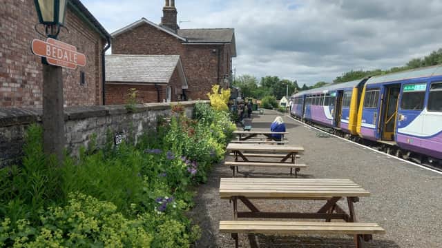 Bedale Station features a sign, flowerbeds, wooden benches, and trains at the platform under a cloudy sky.