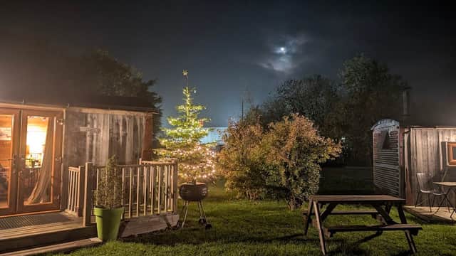 Cozy glamping site at night with a lit Christmas tree, wooden cabins, and a picnic table under a cloudy sky.