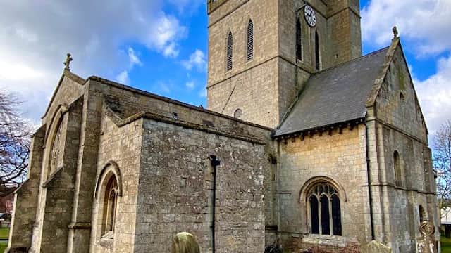 Historic church with a tall clock tower, surrounded by gravestones and lush green grass under a blue sky.