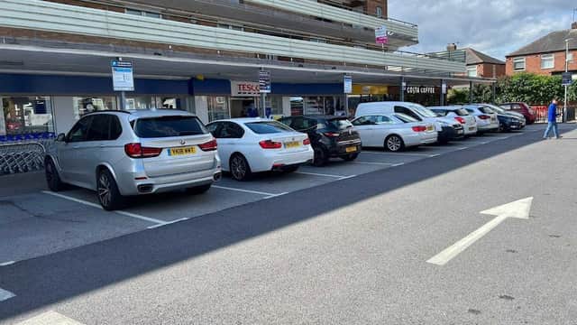 A row of parked cars in a car park with a Tesco store in the background and a cloudy sky above.