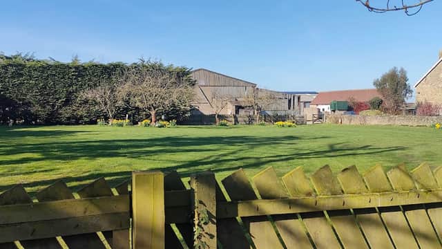 A green field with a wooden fence, surrounded by trees and buildings under a clear blue sky at Colburn Grange Farm.