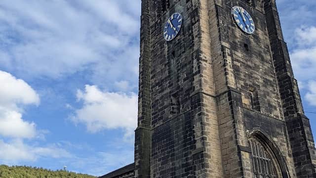 Historic stone clock tower with blue clock faces, surrounded by trees and a partly cloudy sky.