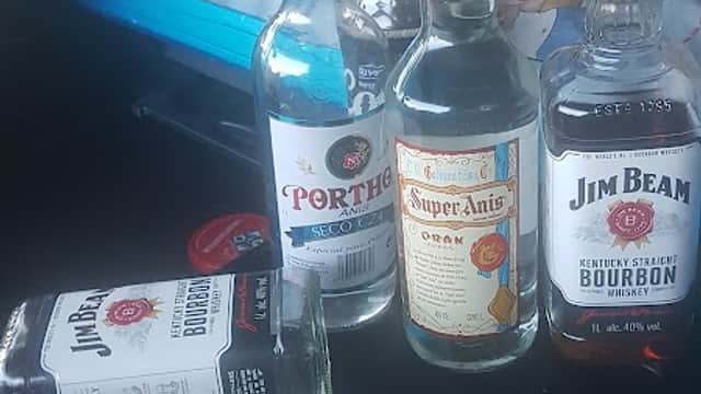Bottles of various spirits and a cup of dark liquid on a table, with a colorful bedspread in the background.