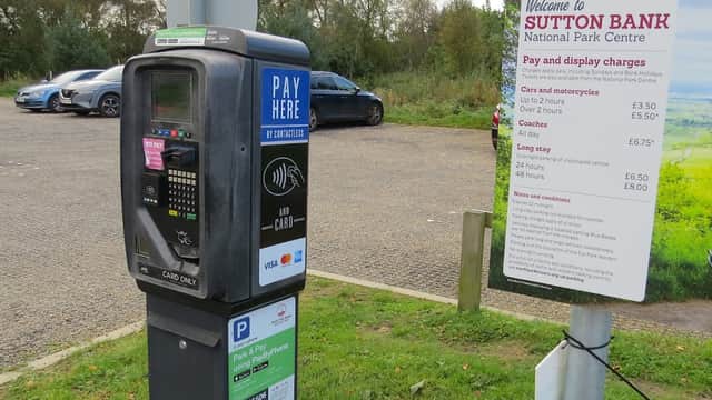 Pay & Display machine and sign detailing parking charges at Sutton Bank Car Park, surrounded by greenery and vehicles.