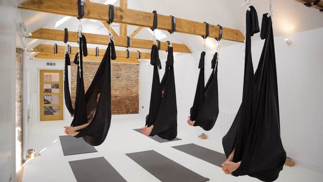 A bright yoga studio with suspended black hammocks, wooden beams, and yoga mats on a white floor.