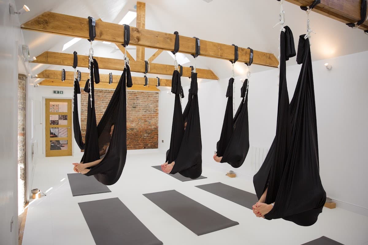 A bright yoga studio with suspended black hammocks, wooden beams, and yoga mats on a white floor.