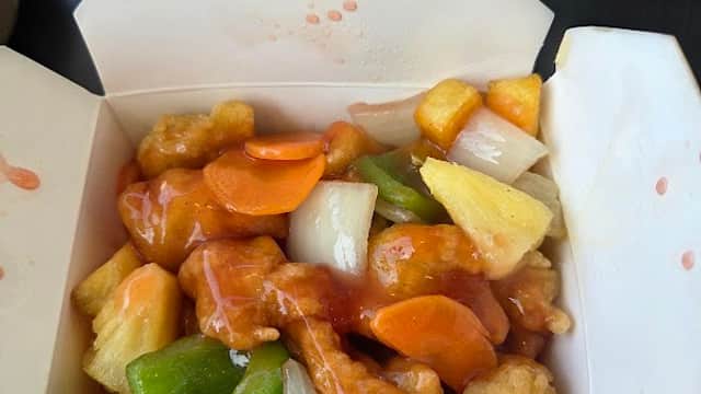 Takeout box filled with sweet and sour chicken, bell peppers, pineapple, and carrots in a tangy sauce.