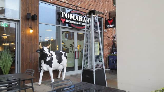 Cow statue outside Tomahawk restaurant, featuring outdoor seating and a patio heater in a brick building.