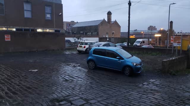 Cobblestone car park with parked cars, buildings in the background, and a cloudy sky in Bradford, Yorkshire.