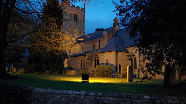 St Michael's church illuminated at dusk, surrounded by trees and gravestones in Kirklington, North Yorkshire.