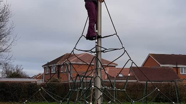Child standing on top of a climbing structure in a playground, giving a thumbs-up, with houses in the background.