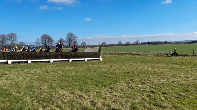 Horses and riders jump a fence in a grassy field under a blue sky, with trees and distant hills in the background.
