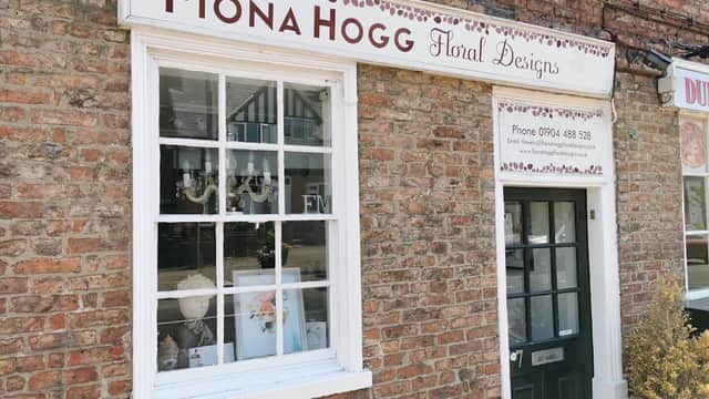 Sign for Fiona Hogg Floral Designs on a brick building, with a window displaying floral arrangements and a door.